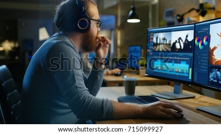 Male Videographer Edits and Cuts Footage and Sound on His Personal Computer, Puts on His Monitors/ Headphones. His Office is Modern and Creative Loft Studio. Royalty-Free Stock Photo #715099927