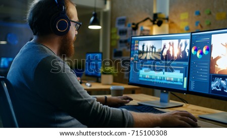 Male Videographer Edits and Cuts Footage and Sound on His Personal Computer, Puts on His Monitors/ Headphones. His Office is Modern and Creative Loft Studio. Royalty-Free Stock Photo #715100308
