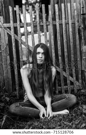 Attractive young long-haired woman in jeans sits near a wooden fence. Contrasting black-and-white photography.