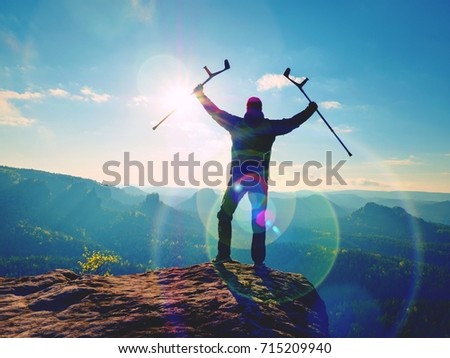 Tourist with medicine crutch above head achieve mountain peak. Hurt backpacker with broken leg in immobilizer stay above valley . The silhouette of man with hand in air
