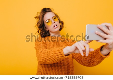 Pretty girl with short curly hair holding smartphone and typing message in front of yellow wall. Fascinating girl with trendy hairstyle wearing sunglasses making selfie on bright background.