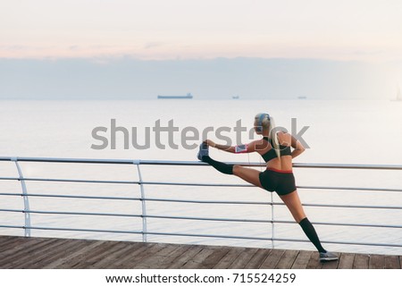 The young beautiful athletic girl with long blond hair in headphones listening to music and doing stretching at sunrise over the sea, back view
