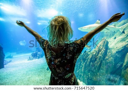 Happy caucasian blonde woman with open arms looking at fishes and shark in oceanarium. Female tourist enjoying in ocean exhibit tank. Lisbon, Portugal. Tourism, holidays, lifestyle and leisure concept Royalty-Free Stock Photo #715647520
