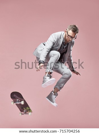 Young handsome serious man with glasses, brown hair and beard, wearing light grey suit and sneakers, jumping with the skateboard on light pink background 
