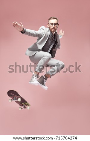 Young handsome serious man with glasses, brown hair and beard, wearing light grey suit and sneakers, jumping with the skateboard and flying in the air on light pink background  Royalty-Free Stock Photo #715704274