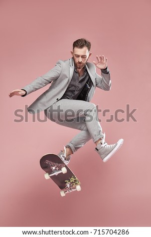 Young handsome man with brown hair and beard, wearing light grey suit and sneakers, jumping with the skateboard on light pink background  Royalty-Free Stock Photo #715704286