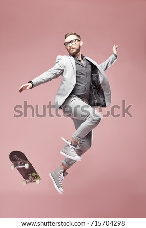 Young handsome funny man with glasses, brown hair and beard, wearing light grey suit and sneakers, jumping with the skateboard on light pink background  Royalty-Free Stock Photo #715704298