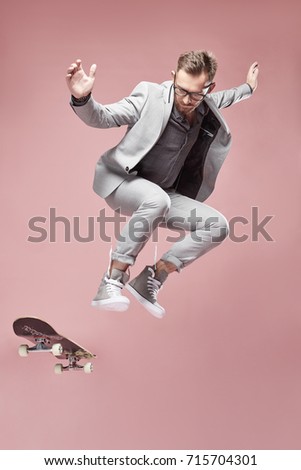 Young handsome serious man with glasses, brown hair and beard, wearing light grey suit and sneakers, jumping with the skateboard and flying on light pink background  Royalty-Free Stock Photo #715704301