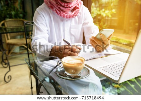 Arab muslim business man ware white traditional clothing in hand writing something on paper and look at smart phone with laptop on coffee glass table