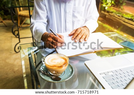 Arab muslim business man ware white traditional clothing and write on paper ,On table have laptop smartphone and cup of coffee