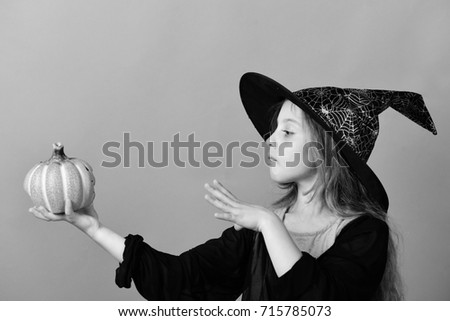 Kid in black witch hat, dress and concentrated face. Girl with carved orange pumpkin isolated on green background. Halloween and autumn concept. Child in witch costume puts spell on jackolantern