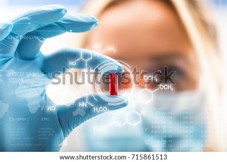 Young attractive female scientist holding a red transparent pill with futuristic scientific air interface with chemical formulas and research data in the foreground Royalty-Free Stock Photo #715861513