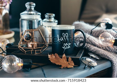Still life details of living room. Cup of coffee on rustic wooden tray, candle and warm woolen sweater on table, decorated with led lights. Autumn weekend concept. Fall home decoration. Royalty-Free Stock Photo #715918423