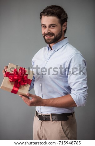 Handsome bearded man in smart casual wear is holding a gift box, looking at camera and smiling, on gray background