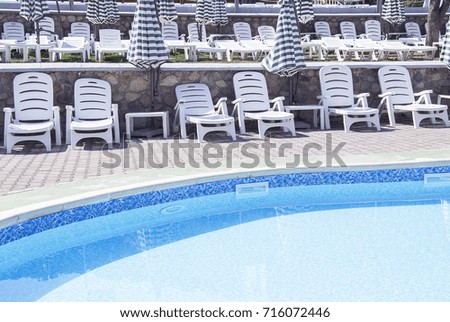 Empty white chaise longues & sun umbrellas near an open air swimming pool with blue water.