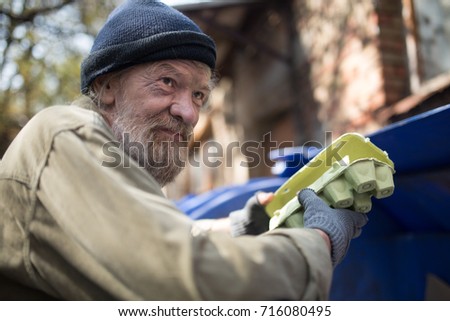 Dirty homeless man holding packing for eggs, standing by the trash can. Lifestyle of tramp, living in the streets. Royalty-Free Stock Photo #716080495