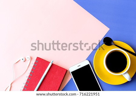 Pastel office desk with smart phone, red notebook, ear phone, spoon and a cup of black coffee on two tone background