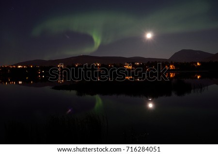 majestic aurora borealis, northern light over calm mirror lake at night with buildings in the background
