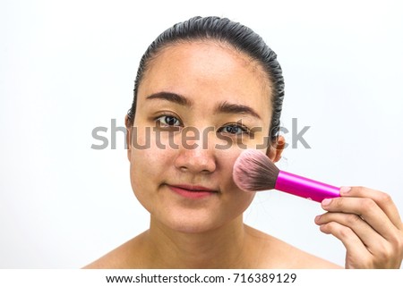 asian woman applying make up with a brush at her face and looking at camera on white background