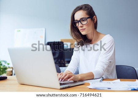 Serious beautiful young woman typing on laptop in a bright modern office Royalty-Free Stock Photo #716494621