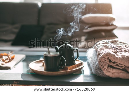 Still life details in home interior of living room. Sweaters and cup of tea with steam on a serving tray on a coffee table. Breakfast over sofa in morning sunlight. Cozy autumn or winter concept. Royalty-Free Stock Photo #716528869