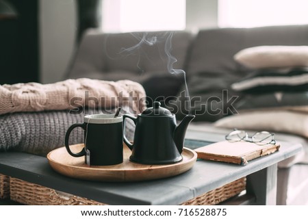 Still life details in home interior of living room. Sweaters and cup of tea with steam on a serving tray on a coffee table. Breakfast over sofa in morning sunlight. Cozy autumn or winter concept. Royalty-Free Stock Photo #716528875