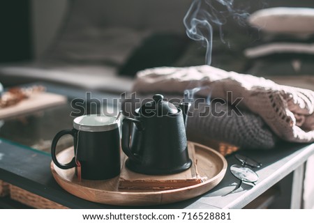 Still life details in home interior of living room. Sweaters and cup of tea with steam on a serving tray on a coffee table. Breakfast over sofa in morning sunlight. Cozy autumn or winter concept. Royalty-Free Stock Photo #716528881
