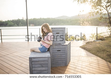 Asian woman is looking to her camera while sitting on the wooden boxes in the beautiful lake scenery view background in the evening sunset
