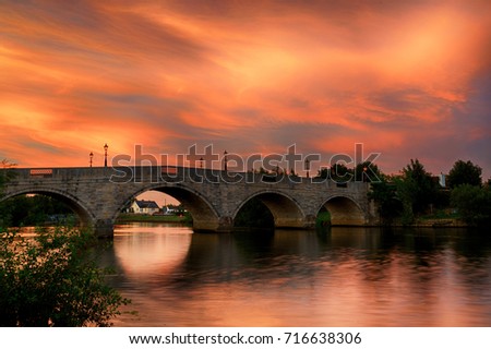 Colorful sky at sunset over Chertsey Bridge, Surrey, UK. Beautiful red, yellow, orange and pink clouds reflected in the River Thames as it flows under the stone built arches of the old bridge. Royalty-Free Stock Photo #716638306