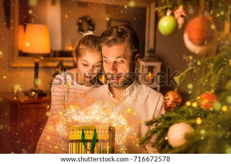 Christmas, family time - caucasian father and daughter looking into a gift (present) bag. Moody warm (gold) light, magical sparkles, cozy atmosphere, by night. Decorated tree and room as background.