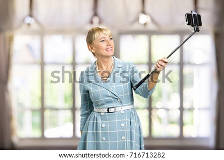 Smiling woman taking selfie with monopod. Attractive mature woman taking selfie with monopod on blurred background. Happy white-skin woman with monopod.