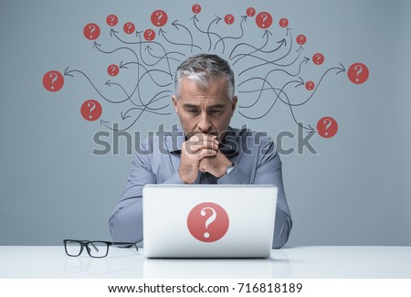 Pensive businessman sitting at desk and working with a laptop, he has question marks and arrows around his head, problem solving and uncertainty concept Royalty-Free Stock Photo #716818189