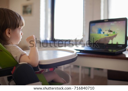A little blond boy watching cartoons on the laptop while sitting on children's chair.