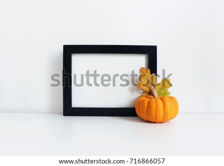 Black blank wooden frame mockup with an orange pumpkin and golden oak leaves lying on the white table. Poster product design. Styled stock feminine photography. Home decor. Autumn, fall concept.