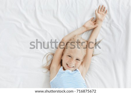Happy lovely blond little female child relaxing in bed, lying on white bedcloth and stretching, enjoying weekend morning, going to watch cartoons on TV and eat breakfast that mother made for her