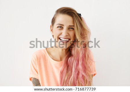 Close up shot of attractive positive young European female with long messy hair with pink highlights enjoying nice day, looking at camera and smiling cheerfully, showing her prefect white teeth