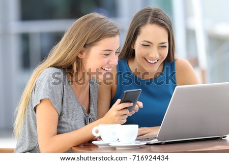 Two happy friends using a laptop and a smart phone sitting in a bar terrace