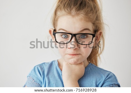 Close up of little cute girl with big blue eyes and light hair looking at camera with angry face expression and raised eyebrows in glasses.