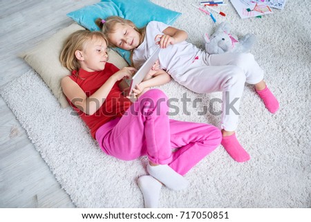 High angle view of pretty little friends watching educational program on digital tablet while lying on cozy carpet at spacious living room