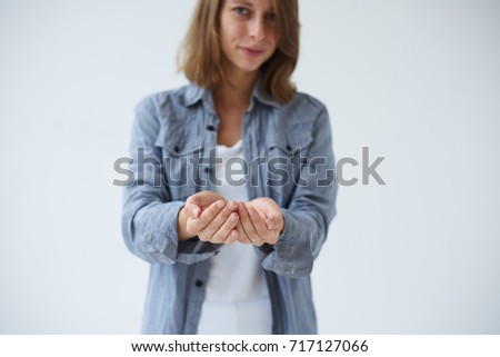 Concept of begging. Cropped studio shot of attractive young Caucasian woman standing at white wall with empty open hands as if holding blank product or object. Signs, gesture and body language.