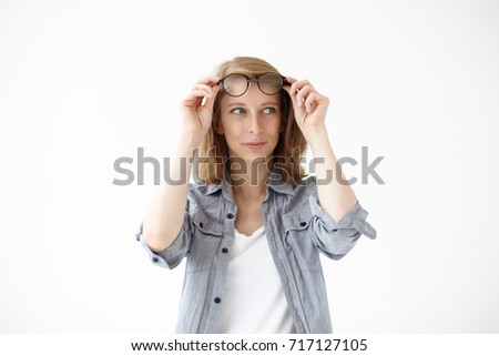 Picture of cheerful young woman dressed casually putting off her stylish eyeglasses in black frame and looking away with curious mysterious expression on her face. People and lifestyle concept.