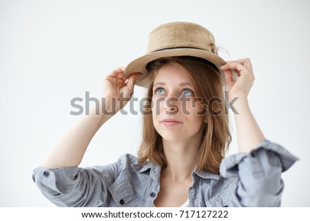 People, clothing, design, style, fashion and shopping concept. Isolated studio picture of gorgeous blue-eyed young woman with freckles trying on vintage round hat while shopping at flea market.