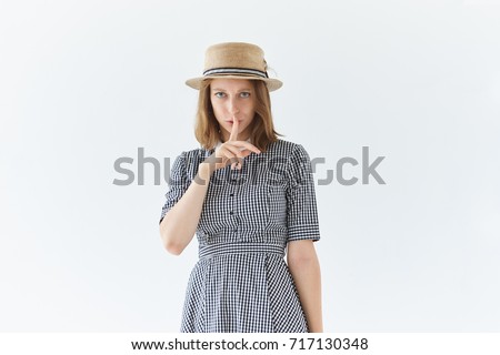 Picture of attractive worried serious young European woman in dress and hat keeping index finger at her lips, saying Shh, Hush, asking to keep silent. Secrecy, silence and conspiracy concept.
