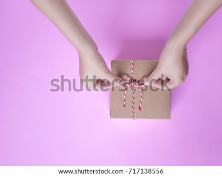 Female hands bow tie a simple craft paper gift box wrapped with colorful rope with as a present for Christmas, new year, valentine day or anniversary on pink background, top view