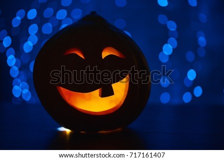 Funny smiling red pumpkin with fire inside and big eyes, prepared for Halloween holiday, sitting on wooden table in dark, against blue lights. Hand made decoration for autumn holiday.