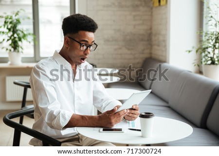 Photo of shocked young african man sitting coworking with tablet computer. Looking aside.