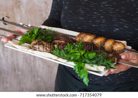 Waiter brings shish-kebab with grilled vegetables on white dish. Meat, field mushrooms and fresh green parsley, barbecue and natural food preparing in restaurant, close up
