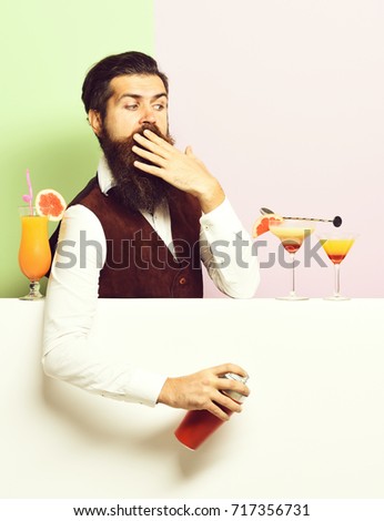 handsome bearded barman with long beard and mustache has stylish hair on surprised face holding shaker and made alcoholic cocktail in vintage suede leather waistcoat on purple green studio background