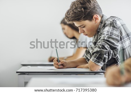 Teenage schoolboy sitting at classroom and doing exam with his classmates. Royalty-Free Stock Photo #717395380