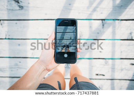 Young social media influencer with marketing ideas makes photo on smartphone of sneaker or running shoes of famous brand, standing on wooden bridge floor